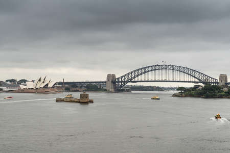 Sydney, Australia - March 21, 2017: Sydney Harbour bridge, Opera House and Denison Fort under gray rainy sky. Darker gray water in front with small boats drawing white lines.のeditorial素材