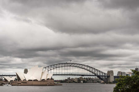 Sydney, Australia - March 23, 2017: White Opera House and black bow of Harbour Bridge behind under heavy rainy cloudscape. Kirribilli tall buildings in back. Gray harbor water up front.のeditorial素材