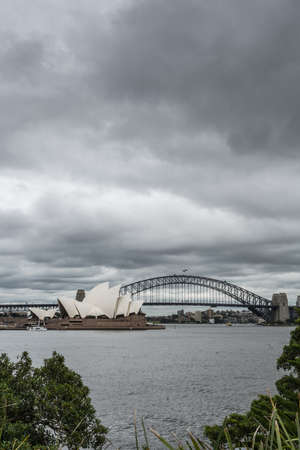 Sydney, Australia - March 23, 2017: Portrait of white Opera House and black bow of Harbour Bridge behind under heavy rainy cloudscape. Gray harbor water and green of Macquarie peninsula in front.のeditorial素材