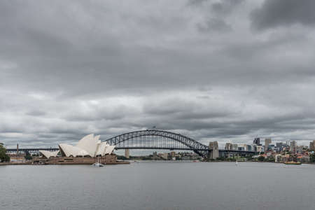 Sydney, Australia - March 23, 2017: White Opera House and black bow of Harbour Bridge under heavy rainy cloudscape. Kirribilli tall buildings in back. Gray harbor water up front, some small boats.のeditorial素材
