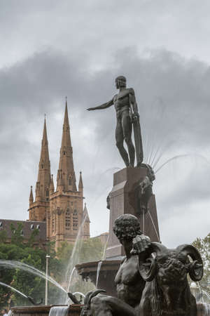 Sydney, Australia - March 23, 2017: Details of black Archibald fountain backed by the two spires of Saint Mary Cathedral, all under gray rainy cloudscape. Some green of surrounding park.のeditorial素材