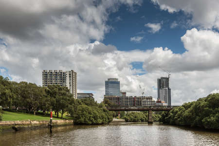 Parramatta, Australia - March 24, 2017: Approaching the city on the river, looking at Macarthur Street bridge and tall office buildings, all under heavy cloudscape. Green shores.のeditorial素材