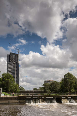 Parramatta, Australia - March 24, 2017: The circular basin is Parramatta harbor. A dam with open floodgates controls flow in river. Heavy cloudscape. Green belt and office tower under construction.のeditorial素材