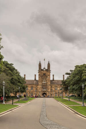 Sydney, Australia - March 25, 2017: Brown stone entrance and clock tower to quadrangle at University of Sydney under heavy sky. Road leading to it, green trees on side.のeditorial素材