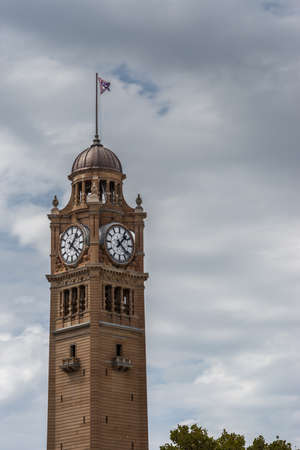 Sydney, Australia - March 25, 2017: Closeup of top of brown stone railway station clock tower against heavy sky. Flag on top.のeditorial素材