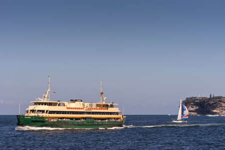 Sydney, Australia - March 26, 2017: Collaroy green-yellow ferry boat in front of outlet to Tasman Sea and South Head cliffs. Small sailing boat. All under open blue sky.のeditorial素材