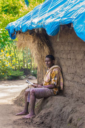 Coorg, India - October 29, 2013: Dubare Elephant Camp. Adult man sits against his dirt hut with straw and blue tarp roof. Holds short club in hand. Green forest background.のeditorial素材