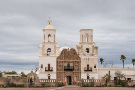 Tucson, Arizona, USA - January 9, 2018: White and brown front facade of Historic San Xavier Del Bac Mission under heavy gray, white cloud deck. Some greenish desert plants in front.のeditorial素材