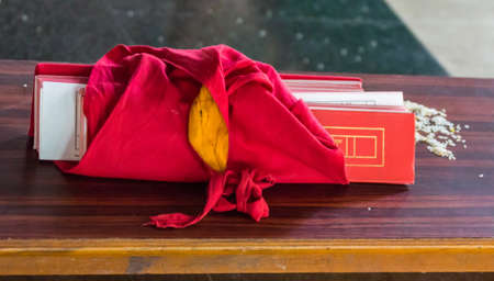 Coorg, India - October 29, 2013: Inside Padmasambhava Vihara of Namdroling Buddhist Monastery. Set of prayer books of monk or nun, captured in red fabric, displayed on bench with rice kernels.のeditorial素材