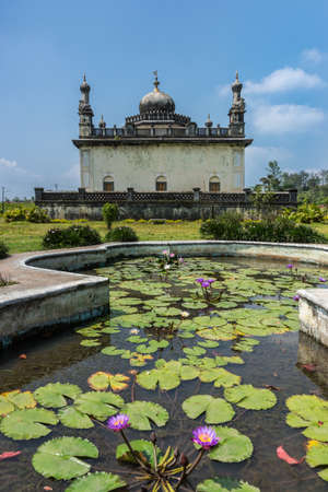 Madikeri, India - October 31, 2013: Royal white and gray mausoleum behind pond with water lilies set in green garden of domain Raja Tombs under blue sky with clouds. Green vegetation.のeditorial素材