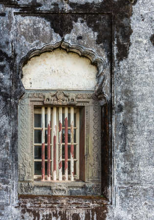 Madikeri, India - October 31, 2013: Closeup of window and rusty decorated metal frame of Royal mausoleum, set in white wall devastated by black mold. Hindu figurines.の写真素材