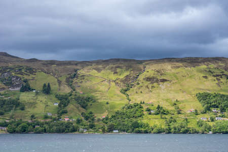 Ullapool, Scotland - June 8, 2012: Hills on west shore of Loch Brrom near Ullapool is green and brown with small white house hidden in green, all under heavy dark cloudscape darkening the water.のeditorial素材