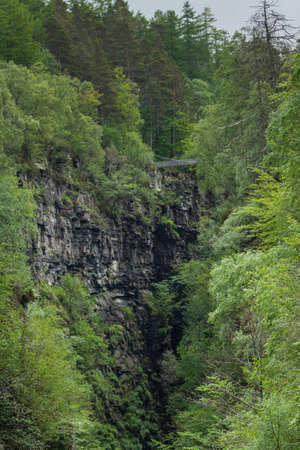 Corrieshalloch Gorge, a deep cut in landscape with forested vertical slopes. Suspension bridge over chasm. Focus on black rocks and abundance of green trees.の写真素材