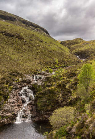 Braemore, Scotland - June 8, 2012: Closeup of Dundonnell Rvier rapids along A832 road offers bronwn rocks and grassy hills under dark cloudscape. Hills in back. Brown pool in front.のeditorial素材