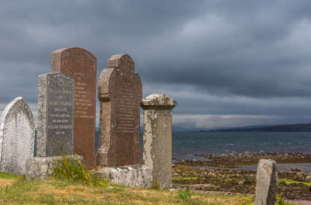 Laide, Scotland - June 8, 2012: Ruins at Laide historic beach side cemetery under dark gray heavy cloudscape. Brown stones and tomb remnants on grass. Gray Atlantic ocean water.のeditorial素材