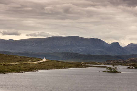 Loch Tollaidh, Scotland - June 9, 2012: Heavy dark brown cloudscape over lake colors water as brown. Set between rocky mountains. Small islets in water. Green grass in front. One-lane road in distance.のeditorial素材