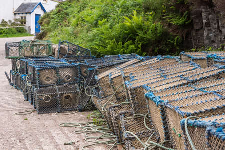 Stromeferry, Scotland - June 10, 2012: Closeup of stack of lobster traps, blue frames and beige nets, on the shore of Loch Carron. Green vegetation and white building.のeditorial素材
