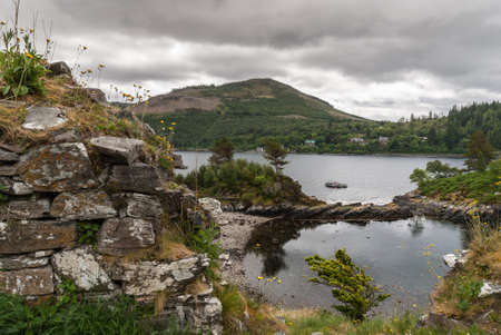 Stromeferry, Scotland - June 10, 2012: Wide shot, pool in rocks at base of Castle Strome ruins on green hill. Cloudscape and motor boat anchored on Loch Carron. Mountains on opposite shore.のeditorial素材
