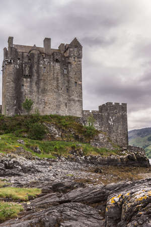Dornie, Scotland - June 10, 2012: Gray-brown-stone main house and tower of Eilean Donan Castle. Green vegetation on top of rocky loch shore up front. Gray cloudscape . Mountains as backdrop.のeditorial素材
