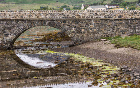 Dornie, Scotland - June 10, 2012: One bow of footbridge to Eilean Donan Castle. Half reflected on waters during low tide. Green hill with houses as backdrop.のeditorial素材