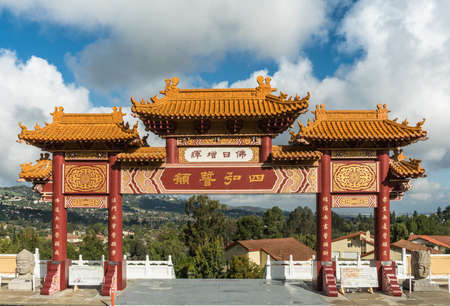Hacienda Heights, CA, USA - March 23, 2018: Red and ochre colored Torii enrance gate to Hsi Lai Buddhist Temple under blue-white cloudscape. Mandarin characters sign. Green hills in distance.のeditorial素材
