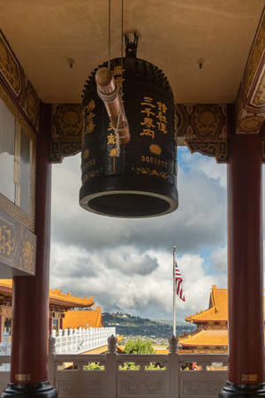 Hacienda Heights, CA, USA - March 23, 2018: Giant ceremonial bell hinging in front of main shrine of Hsi Lai Buddhist Temple. cloudscape, American flag, ochre roofs and green hills on horizon.のeditorial素材