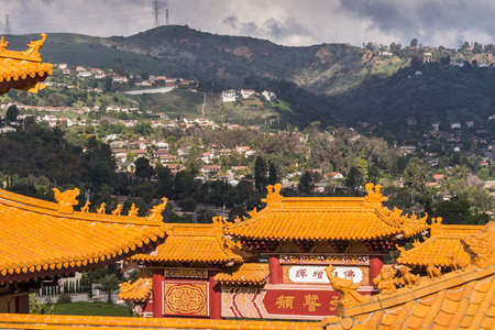Hacienda Heights, CA, USA - March 23, 2018: Red ochre roof structures of Hsi Lai Buddhist Temple under dark heavy cloudscape with green and dark hills and housing in the background.のeditorial素材