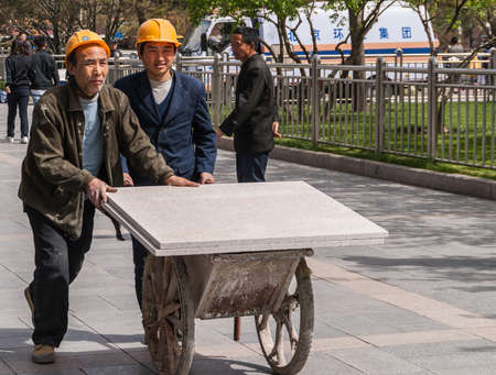 Beijing, China - April 27, 2010: Two workmen with yellow helm and in work garb push cart with large white stone slabs at Tienanmen Square.  Other people, a van and park green.のeditorial素材