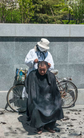 Beijing, China - April 29, 2010: Street scene where ambulant barber with his bike cuts hair of black covered man. Gray wall of green park as background. Cut black hair on sidewalk.のeditorial素材