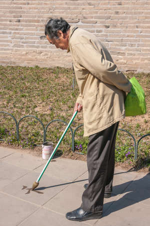 Beijing, China - April 29, 2010: Temple of Heaven park. Man with beige coat engaged in calligraphy uses larg pencil-brush to design Mandarin character on sidewalk surface using water.のeditorial素材