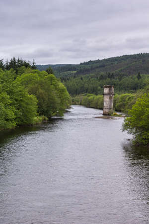 Fort Augustus, Scotland - June 11, 2012: Brown remnant of tower of disappeared bridge over silver Oich River, flanked by green forests along its shores under heavy gray sky.のeditorial素材