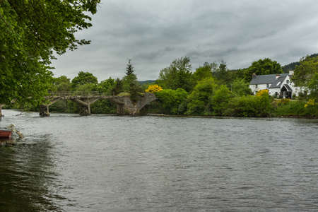 Fort Augustus, Scotland - June 11, 2012: Damaged wooden foot bridge over River Oich. Green belt between silver water and heavy gray cloudscape. White cottage in photo.のeditorial素材