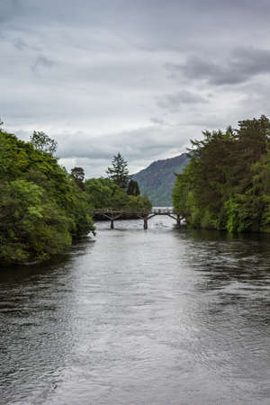 Fort Augustus, Scotland - June 11, 2012: Damaged wooden foot bridge over River Oich. Green belt between silver water and heavy gray cloudscape. Hills on horizon.のeditorial素材