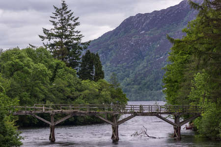 Fort Augustus, Scotland - June 11, 2012: Closeup of Damaged wooden foot bridge over River Oich. Green belt between silver water and heavy gray cloudscape. Hills on horizon.のeditorial素材