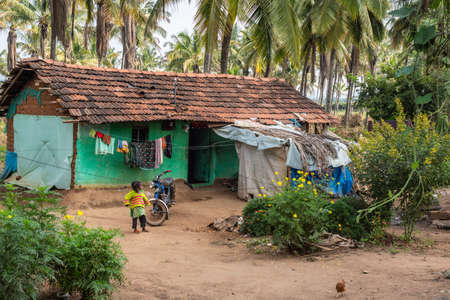 Belathur, Karnataka, India - November 1, 2013: Little girl plays in front of green peinted humble dwelling with red pane roof. Green palm trees in back. flowers, dirt, laundry and chickens.のeditorial素材
