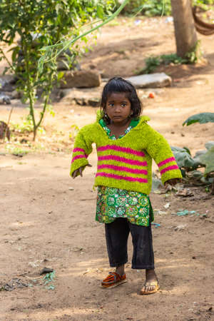 Belathur, Karnataka, India - November 1, 2013: Closeup of young girl with black hair and wide black eyes, wearing too large yellow-pink vest playing in the dirt garden. Different shoes.のeditorial素材