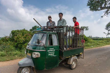 Belathur, Karnataka, India - November 1, 2013: Dark green tricycle car brings four farm workers, smiling men, standing on loading area of car to the field. Road scene with blue sky and white clouds.のeditorial素材