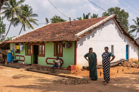Nilavagilukaval, Karnataka, India - November 1, 2013: Two smiling woman and a man in front of their one story stone building with red pan roof. Brown dirt road and open sewer.のeditorial素材
