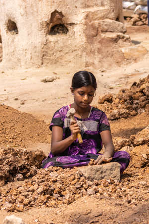 Hunsur, Karnataka, India - November 1, 2013: Seated young girl crushes limestone with heavy hammer. Heap of stones on her side. Part of kiln in back.のeditorial素材