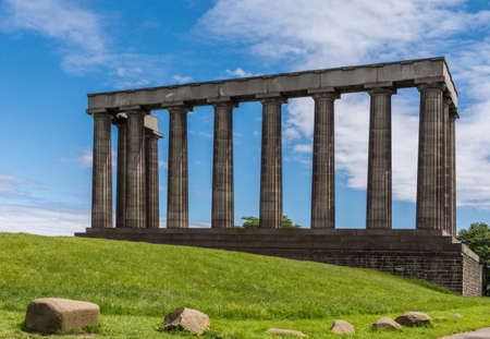 Edinburgh, Scotland, UK - June 13, 2012: National Monument is series of dark brown pilars against blue sky with white clouds. Green grass up front.のeditorial素材