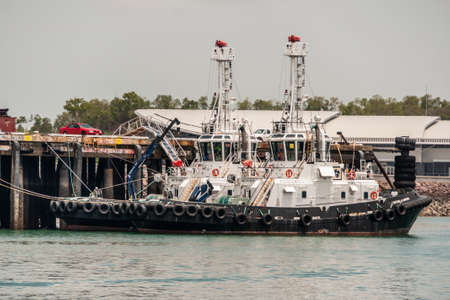 Darwin, Northen Territory, Australia - December 1, 2009: Closeup of two black and white tug boats docked in port under gray sky. Cars onto pier.のeditorial素材