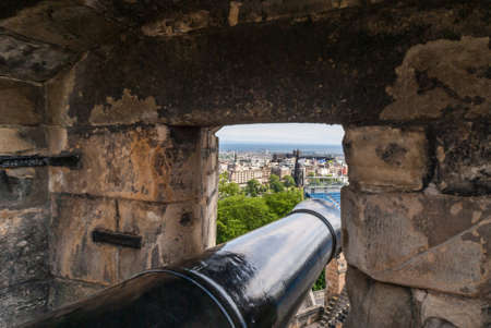 Edinburgh, Scotland, UK - June 14, 2012: Looking along barrel of black cannon set in niche over the city behind gray stone rampart. Black Scott monument spire behind green park. Princess street views.のeditorial素材