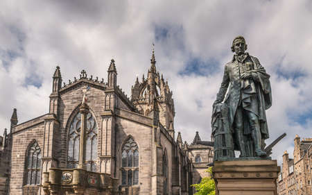 Edinburgh, Scotland, UK - June 14, 2012: Adam Smith bronze statue on market square in front of brown stone Saint Gilles Cathedral crown tower under gray silver sky with blue patches. Mercat Cross pillar.のeditorial素材