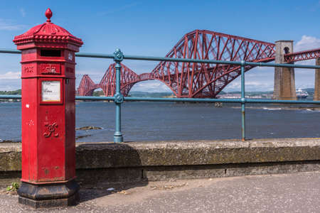 Queensferry, Scotland, UK - June 14, 2012: Closeup of Red metal iconic Forth Bridge for trains over Firth of Forth between blue sky and blue water. Combination with Red Postal mail box on boardwalk.のeditorial素材