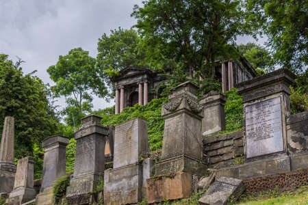 Glasgow, Scotland, UK - June 17, 2012: Necropolis. Shot up the hill of row of molded brown stone tombstones among green foliage. Gray sky. Pillared temple-like structure on top.のeditorial素材