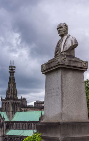 Glasgow, Scotland, UK - June 17, 2012: Necropolis. Gray stone Ralph Wardlaw tombstone with his bust and the Cathedral spire and green roofs in the back under cloudscape.のeditorial素材