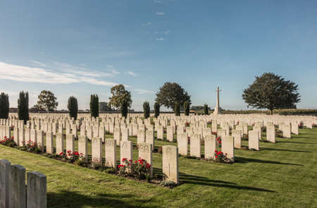 Proven, Flanders, Belgium - September 15, 2018: Overview of Mendinghem British war cemetery under blue morning sky. Green lawn, beige tomb stones and red roses with dark green trees sprinkled.のeditorial素材