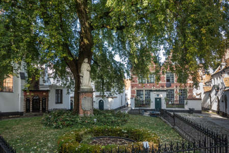 Kortrijk, Flanders, Belgium - September 17, 2018: White Johanna of Constantinople statue under green tree in beguinage courtyard. Small houses around.のeditorial素材