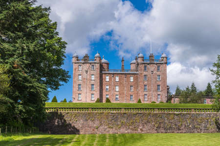 Holm of Drumlanrig, Scotland, UK - June 18, 2012: The side of free standing pink sand stone Drumlanrig Castle on its higher plateau under blue cloudy sky. Green lawn and trees up front.のeditorial素材