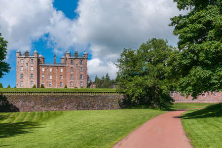 Holm of Drumlanrig, Scotland, UK - June 18, 2012: Long shot along driveway to side of free standing pink sand stone Drumlanrig Castle  under cloudy sky. Green trees and lawns.のeditorial素材
